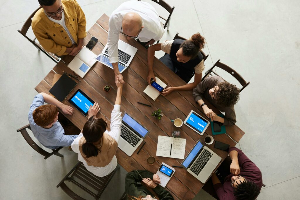 A group of people sit around a table working on various devices including tablets and laptops. They appear to be collaborating on a project.